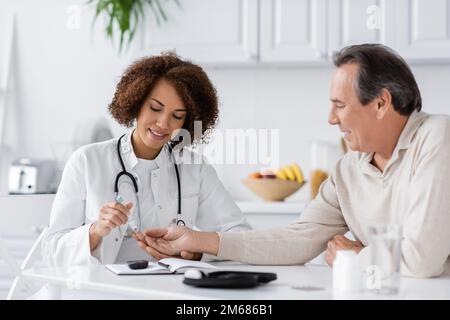 sourire afro-américain médecin prenant un échantillon de sang de patient mature avec lancet stylo, image de stock Banque D'Images