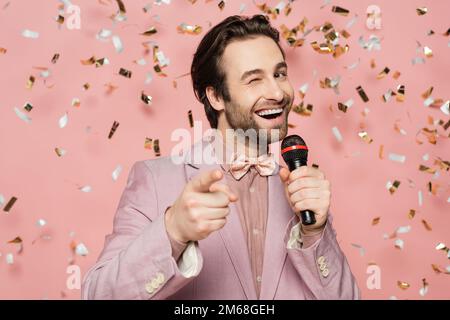 Hôte positif de l'événement tenant le microphone et wencant à la caméra sous confetti sur fond rose, image de stock Banque D'Images