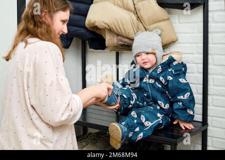 La mère met des chaussures bleues sur le pied de bébé enfant assis dans le couloir de la maison. Femme maman vêtue chaud bottes vêtements sur enfant pour l'hiver marcher dans c Banque D'Images