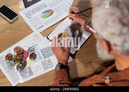 KIEV, UKRAINE - 17 NOVEMBRE 2022 : homme mûr flou avec écriture en bitcoin doré dans un carnet proche de documents et smartphone avec écran vierge Banque D'Images