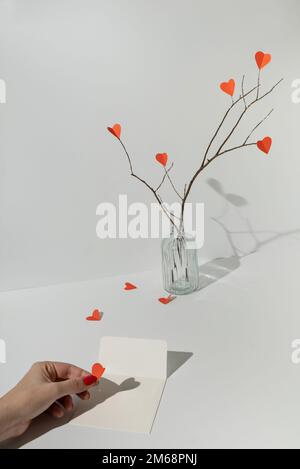 Branches avec coeurs en papier rouge et main femelle mettant le coeur dans une enveloppe blanche. Un minimum d'amour ou de valentine concept. Banque D'Images