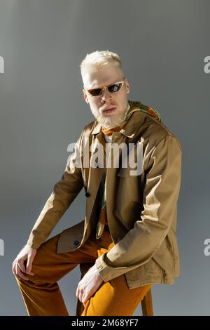 albinos barbu dans des lunettes de soleil élégantes et veste de chemise assis sur chaise haute sur gris, image de stock Banque D'Images