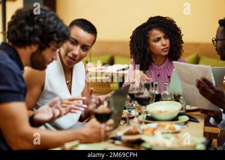Un groupe de gestionnaires multiculturels est assis dans un restaurant à une table, parle d'un projet et utilise les technologies pour l'améliorer. Banque D'Images