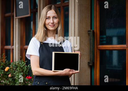 Jeune femme caucasienne de 30 ans avec cheveux blonds courts tenant une pancarte avec espace de copie près des portes du café. Rouvrir le restaurant après le coronavirus Banque D'Images