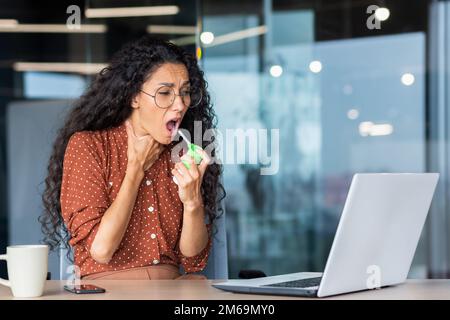 Femme hispanique malade au bureau, femme d'affaires a mal à la gorge, mal à la gorge, utilise des médicaments pour soulager la douleur tout en étant assis au travail à l'intérieur du bureau avec un ordinateur portable au travail. Banque D'Images