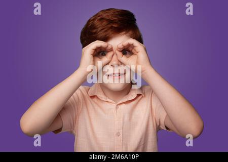 Enfant curieux explorant le monde regardant à travers les doigts en forme de jumelles et exprimant l'étonnement isolé sur fond violet. Banque D'Images