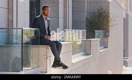 Homme d'affaires ethnique afro-américain responsable indépendant assis à l'extérieur dactylographiant sur un ordinateur portable inquiet au sujet de la problématique homme de travail pensant Banque D'Images