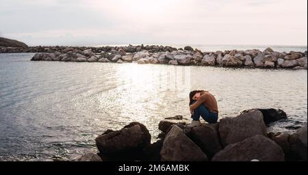 femme assise sur des pierres en face de la mer embrassant ses jambes au coucher du soleil Banque D'Images