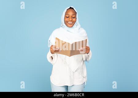 Bonne femme noire dans hijab posant avec un livre ouvert, des recettes de lecture ou des notes sur fond bleu studio, espace libre Banque D'Images