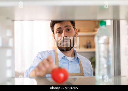 Triste adulte caucasien barbu gars en tablier ouvre la porte du réfrigérateur vide, prend une tomate dans la cuisine intérieure Banque D'Images