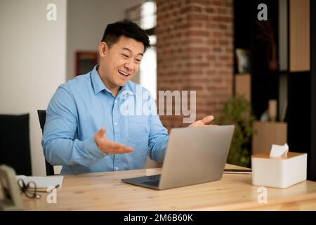 Homme asiatique mature et enthousiaste utilisant un ordinateur portable moderne, des appels vidéo et des gestations à la webcam, assis sur le lieu de travail à la maison Banque D'Images