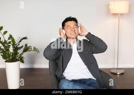 Un homme japonais adulte souriant avec des yeux fermés dans un casque sans fil écoute de la musique, aime la musique dans le salon Banque D'Images