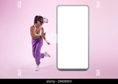 Femme afro-américaine sportive expérimentant le métaverse dans des lunettes de réalité virtuelle près d'un grand smartphone avec écran vierge Banque D'Images