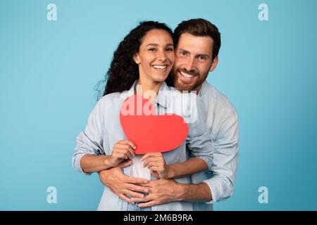 Jeune homme européen souriant en femme hug décontractée et tenant le coeur, isolé sur fond bleu, studio Banque D'Images