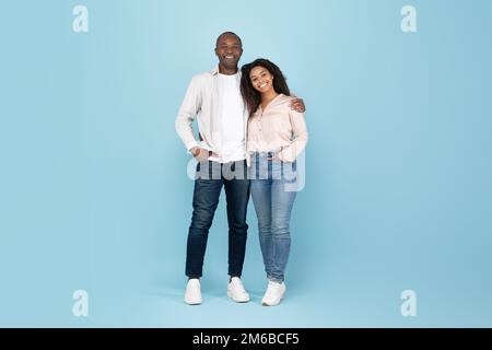 Portrait d'un couple adulte noir heureux qui embrasse, regarde l'appareil photo et sourit, posant sur le mur bleu du studio, sur toute la longueur Banque D'Images