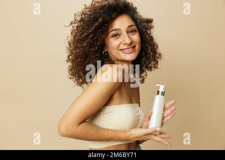 Femme applique de la crème et du baume à ses cheveux bouclés, le concept de protection et de soin avec des produits de salon, un look sain, un sourire aux dents sur un beige Banque D'Images