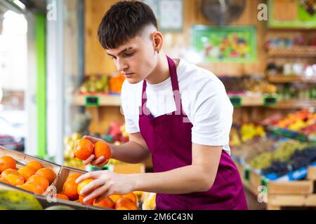 Professionnel jeune homme ouvrier de supermarché en tablier vert arranger des tangerines dans le supermarché Banque D'Images