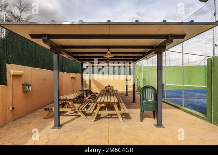 Un pavillon de jardin avec cadre en métal et en bois avec de longues tables en bois donnant sur un court de tennis avec un sol peint en intérieur bleu Banque D'Images