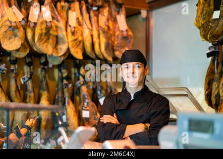 Jeune vendeur d'atelier de boucher en uniforme noir debout avec bras croisés près de la crémaillère avec pattes de jamon suspendues Banque D'Images