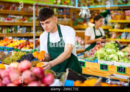 Professionnel jeune homme ouvrier de supermarché en tablier vert arranger des tangerines dans le supermarché Banque D'Images