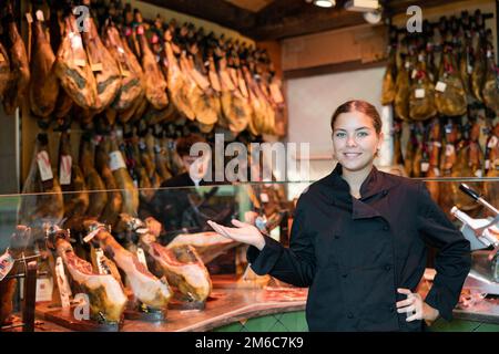 Une salesgieuse souriante en uniforme noir recommandant du jambon ibérique dans un magasin de viande Banque D'Images