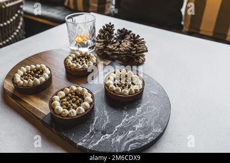 Quatre tartes décorées de crème blanche et de perles dorées placées en deux lignes sur un tableau bicolore. Un porte-bougie et quatre cônes placés à côté de la trancheuse. Coussins flous en arrière-plan. . Photo de haute qualité Banque D'Images
