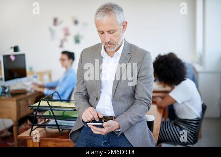 Envoi de mises à jour client. homme d'affaires mature utilisant un téléphone portable tout en étant assis sur une table dans un bureau. Banque D'Images