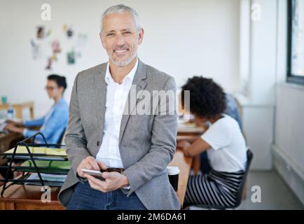 Je garde mes clients au courant. Portrait d'un homme d'affaires mature utilisant un téléphone portable tout en étant assis sur une table dans un bureau. Banque D'Images