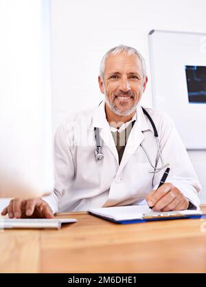 Rester à jour par le biais de revues médicales en ligne. Portrait d'un médecin homme mature travaillant sur un ordinateur de bureau dans son bureau. Banque D'Images