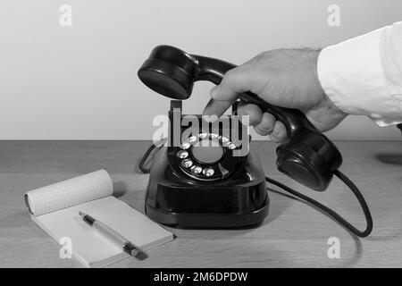 Ancien téléphone installé sur une table en bois, la main de l'homme compose un numéro de téléphone sur un disque téléphonique. installation rétro. Noir et blanc Banque D'Images