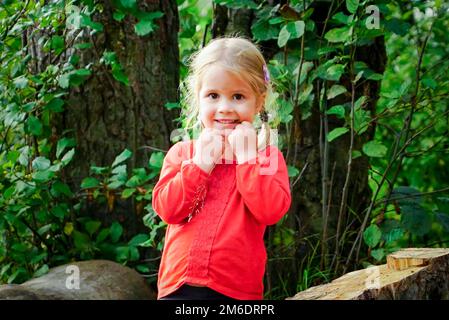 Une petite fille gaie de 3 ans se tient près d'une bûche dans la forêt. Banque D'Images