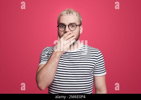 Portrait d'un beau homme excité portant des lunettes regardant l'appareil-photo tout en couvrant les mains de la bouche isolée sur fond rose studio. Banque D'Images