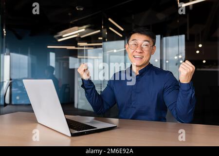 Homme d'affaires asiatique couronné de succès, portrait de l'homme au bureau en chemise décontractée, employé souriant et regardant l'appareil photo satisfait de la réussite en tenant le geste de la victoire main vers le haut. Banque D'Images