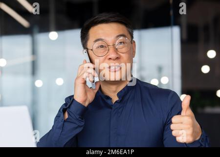 Homme d'affaires asiatique joyeux et réussi parlant au téléphone avec des clients à l'intérieur du bureau, homme mature dans une chemise travaillant sur un ordinateur portable sur le lieu de travail. Banque D'Images