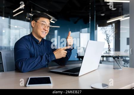 Appel vidéo Happy asian parlant à distance avec des collègues, homme d'affaires au bureau avec ordinateur portable lors d'une réunion en ligne avec les employés, assis au bureau, homme en chemise. Banque D'Images