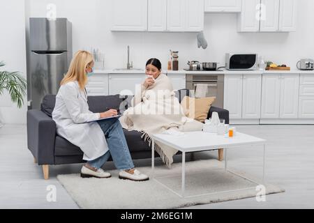 Femme asiatique dans une couverture tenant la serviette près du médecin dans l'écriture de masque médical sur le presse-papiers à la maison, image de stock Banque D'Images