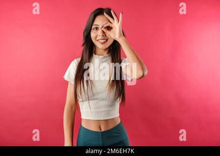 Bonne femme brune portant un haut décontracté isolé sur fond rouge regardant à travers le signe d'okey. Heureux, confiant et tout va bien. Face à l'expressi Banque D'Images