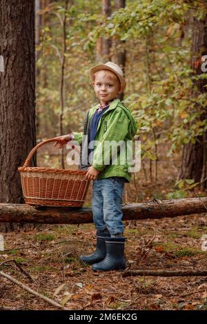 Petit garçon dans un chapeau dans la forêt d'automne. Garçon avec un panier dans les bois. Petit forestier. Près d'un arbre tombé. Banque D'Images