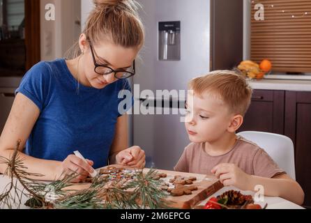 Une jeune mère et un jeune fils en cuisine font des biscuits. Noël pain d'épice homme givrant tuyauterie décoration. Femme avec son enfant décorati Banque D'Images