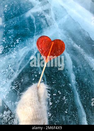 Le lollipop rouge en forme de coeur tient une main dans une mitaine contre le fond de glace. Banque D'Images