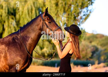 Une fille dans un chapeau noir dans les rayons de le soleil couchant regarde avec joie un cheval Banque D'Images