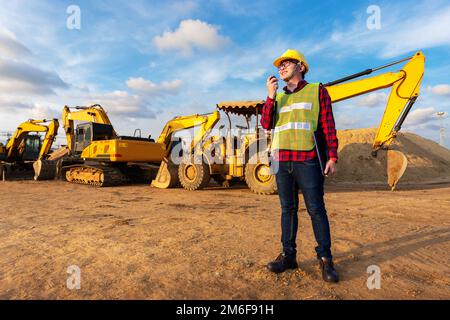 un technicien asiatique, ingénieur civil, parle avec l'équipe par walkie talkie tenir un ordinateur portable ou une tablette pour le transport de travaux de construction avec une pelle hydraulique Banque D'Images