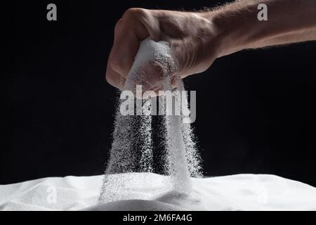 Homme avec une poignée de sable blanc sec dans ses mains, répande du sable à travers les doigts sur fond noir. Concept de flux de vie. Gros plan de grains de pur Banque D'Images