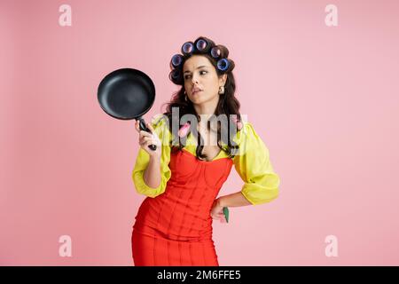 femme de ménage ennuyé dans les cheveux curlers et corset rouge robe regardant la poêle isolée sur rose, image de stock Banque D'Images