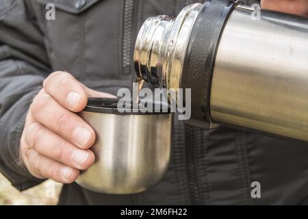 Un homme s'assoit dans une forêt d'automne ou dans un parc et verse le thé d'un thermos dans une tasse, en gros plan Banque D'Images