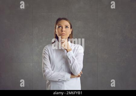 Jeune femme caucasienne pensant et touchant le menton regardant debout sur le mur gris Banque D'Images