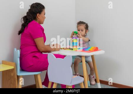 Médecin pédiatre Latina, assis à une table avec une petite fille, jouant dans la salle de jeux de son bureau médical. Banque D'Images