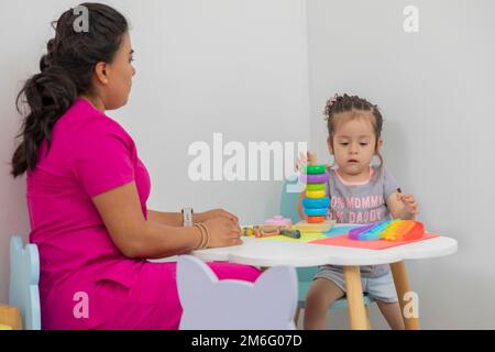 Médecin pédiatre Latina, assis à une table avec une petite fille, jouant dans la salle de jeux de son bureau médical. Banque D'Images