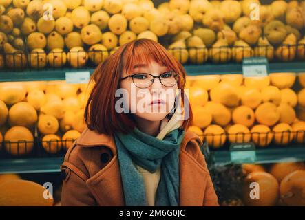 Portrait d'une belle femme au gingembre asiatique, portant un manteau beige et une écharpe bleue sur fond de fruits frais dans une épicerie. Citrons et santé Banque D'Images