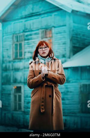 Un magnifique étudiant de gingembre asiatique dans un manteau beige et un foulard bleu gèle un jour froid de janvier sur le fond d'un village en bois bleu ho Banque D'Images